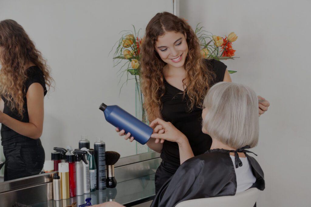Een glimlachende jonge kapster met krullend haar houdt een blauwe fles haarverzorgingsproduct vast terwijl ze een oudere vrouw met zilvergrijs haar adviseert die in een salonstoel zit.