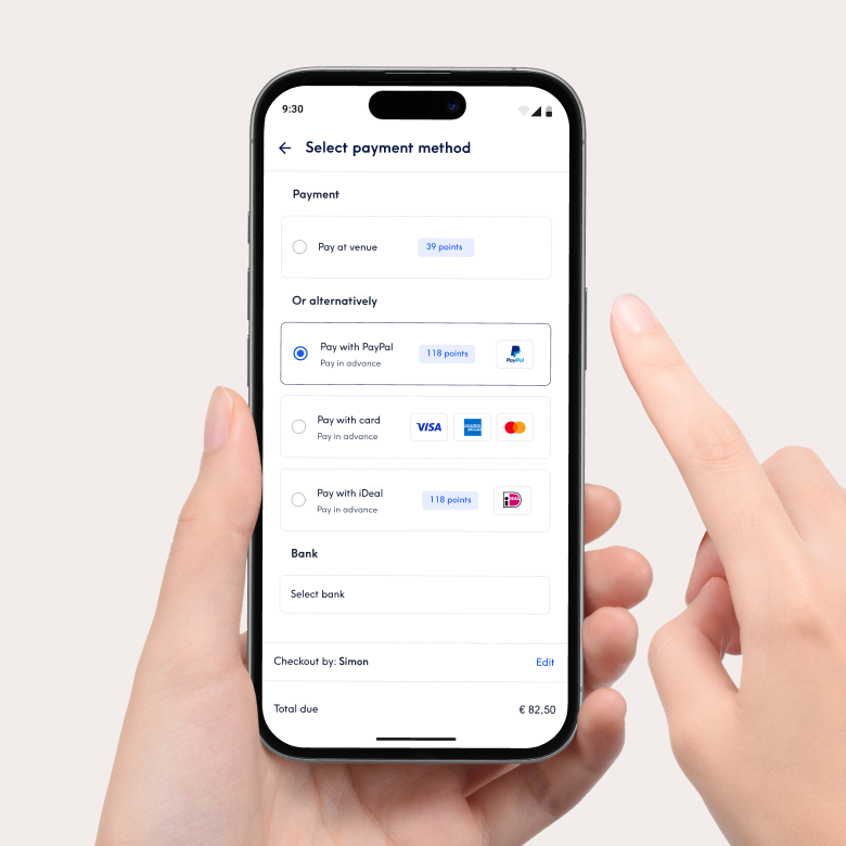 Close-up van een smartphone met de betaalinterface van de Treatwell-app om een betaalmethode te selecteren. Opties zijn onder meer betalen in de salon of vooraf betalen met PayPal, kaart of iDeal. Een hand houdt de telefoon vast und een vinger wijst naar het scherm.