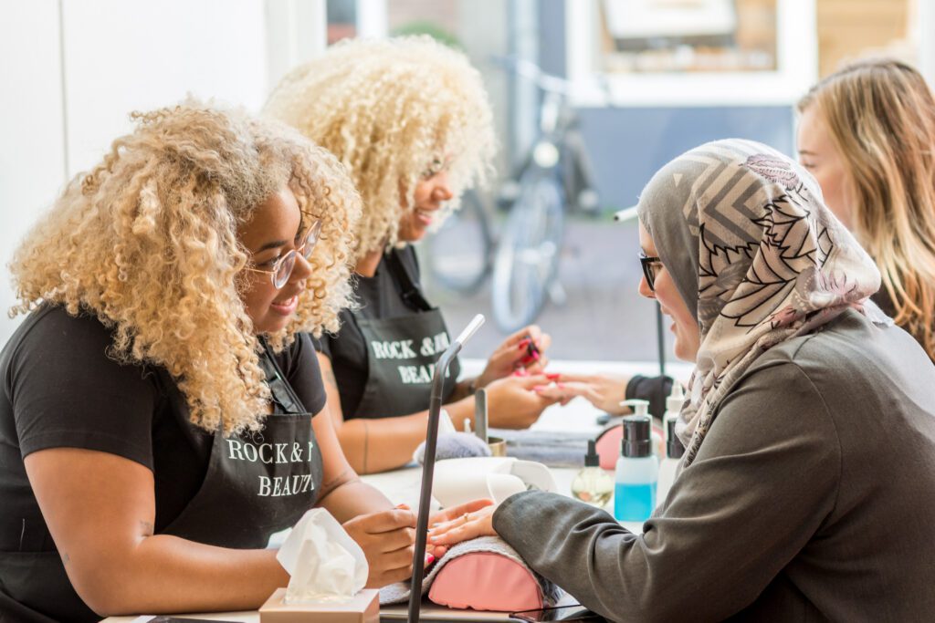Twee manicures met blond krullend haar en zwarte schorten met de tekst "Rock & Roll Beauty" geven manicures aan klanten aan een lange tafel. Eén klant draagt een grijze hidjab.
