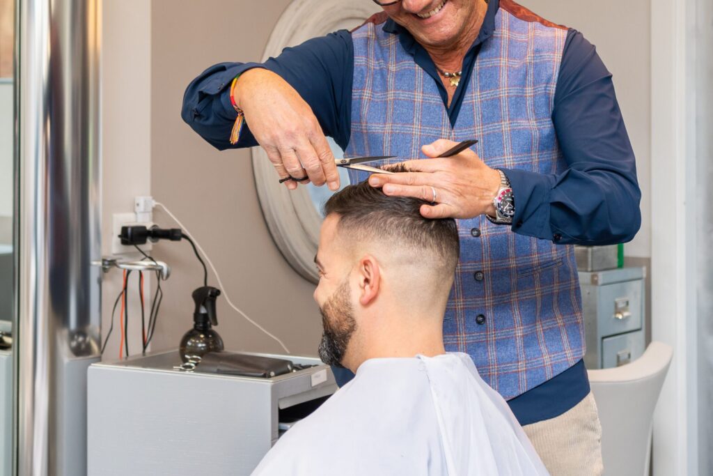 Een kapper met een blauw geruit gilet knipt het haar van een klant met een schaar en een kam in een salon. De klant heeft kort haar, een baard en draagt een witte kappersmantel.