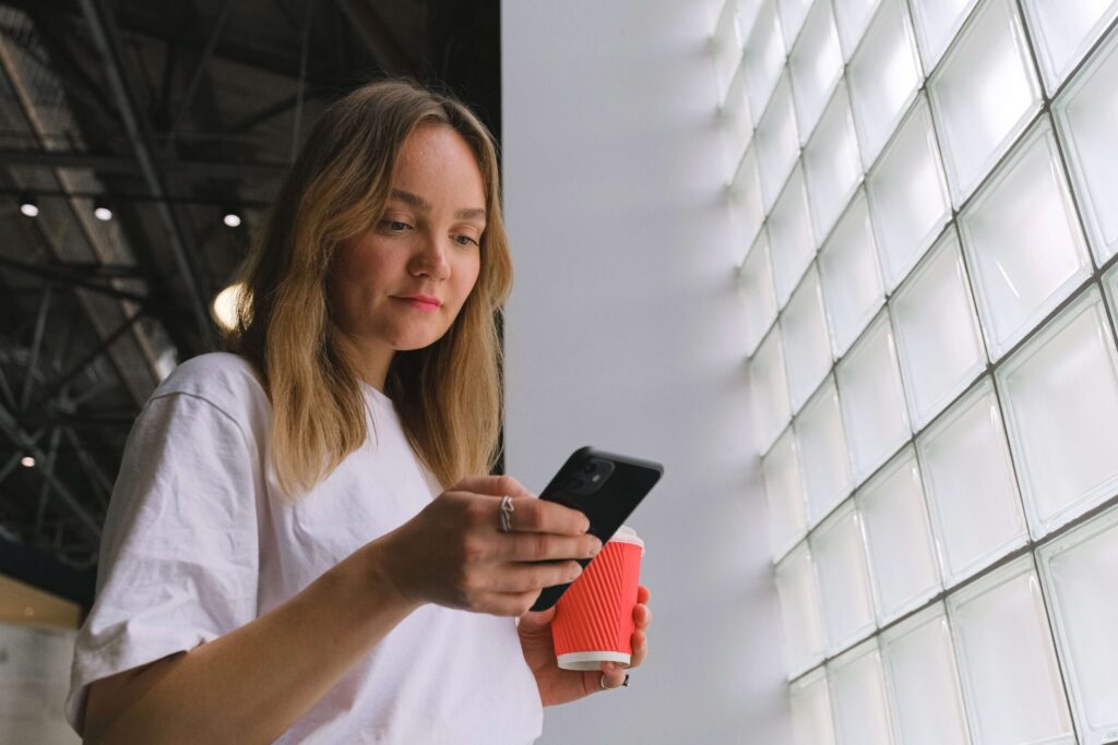 Een kikvorsperspectief van een jonge vrouw met blond haar en een wit t-shirt, die naar haar zwarte smartphone kijkt terwijl ze een rode meeneemkoffiebeker vasthoudt. Ze staat naast een grote muur van glasbouwstenen.