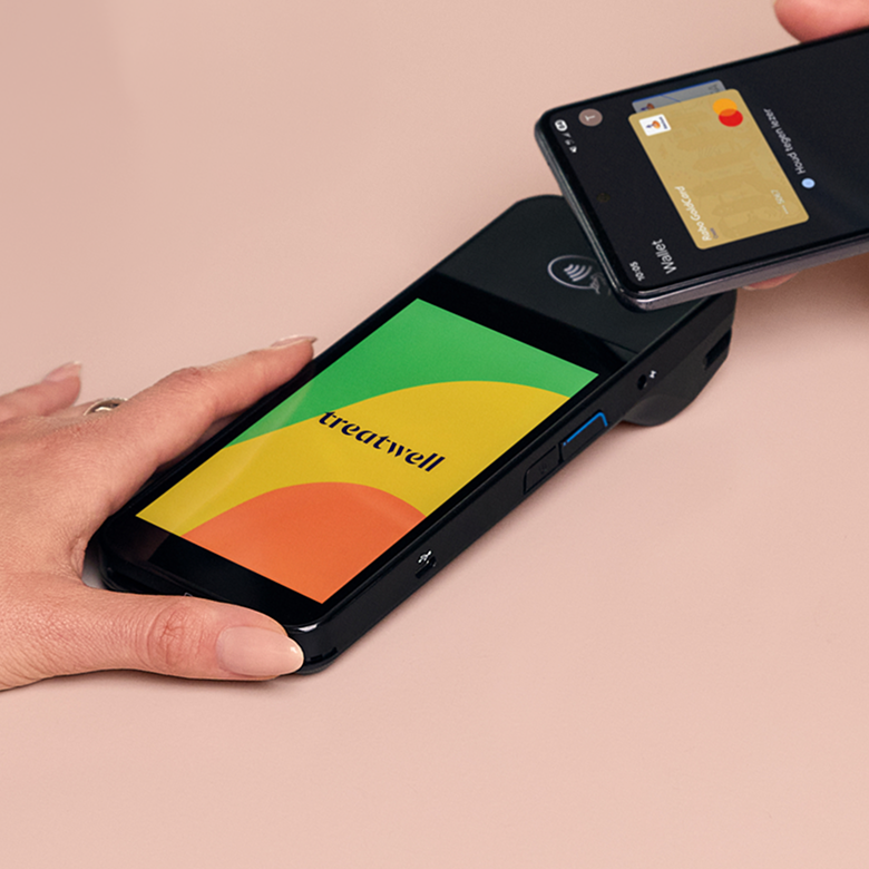 Een persoon houdt een zwarte betaalterminal vast met een meerkleurig scherm met een groen, geel en oranje ontwerp en het donkerblauwe "Treatwell"-logo, terwijl een andere persoon diens smartphone ertegenaan houdt om een contactloze betaling te doen via een digitale portemonnee. De scène speelt zich af op een lichtroze oppervlak.