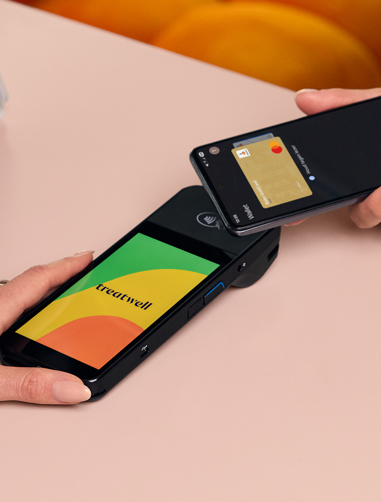 Een persoon houdt een zwarte betaalterminal vast met het lichtblauwe scherm van het merk "Treatwell", terwijl een andere persoon diens smartphone ertegenaan houdt om een contactloze betaling te doen via een digitale portemonnee. De scène speelt zich af op een lichtroze tafel.